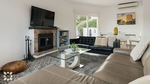 A living room with a fireplace, a black leather sofa, a glass coffee table, and a television mounted above the fireplace. - Villa MONTE CRISTO by Abahana Villas (Teulada)