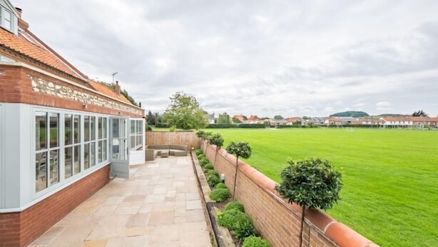A patio area with a brick wall, a windowed section, and a green lawn in the background. - Field Piece Cottage, Burnham Market, Norfolk (Burnham Market)