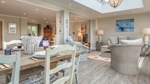 A spacious living area with a high ceiling, a dining table set with plates and glasses, and a comfortable seating area with cushions and a wicker chair. - Field Piece Cottage, Burnham Market, Norfolk (Burnham Market)