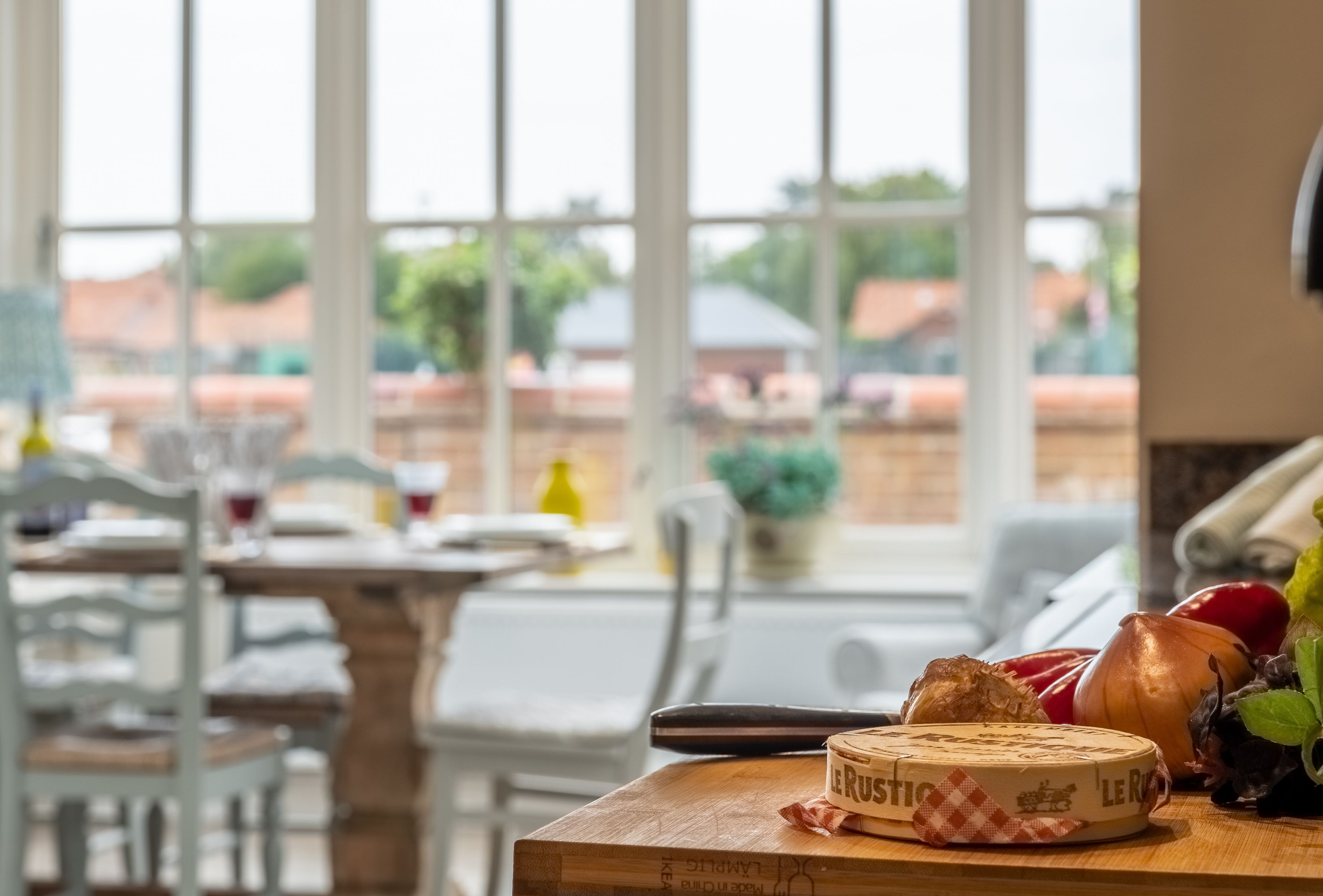 A dining area with a table set for two, a cheese board with a piece of cheese labeled "Le RUSTIC," and a window with a view of buildings and trees.