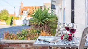 Une table avec un livre et deux verres de vin rouge, le tout placé devant un bâtiment et une végétation luxuriante.