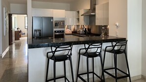 Una cocina moderna con una isla central, taburetes de bar, electrodomésticos de acero inoxidable y vistas al área de estar contigua.