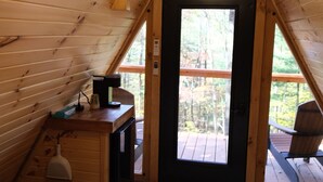 A wooden cabin with a loft bedroom, a desk with a lamp, and a view of the forest.