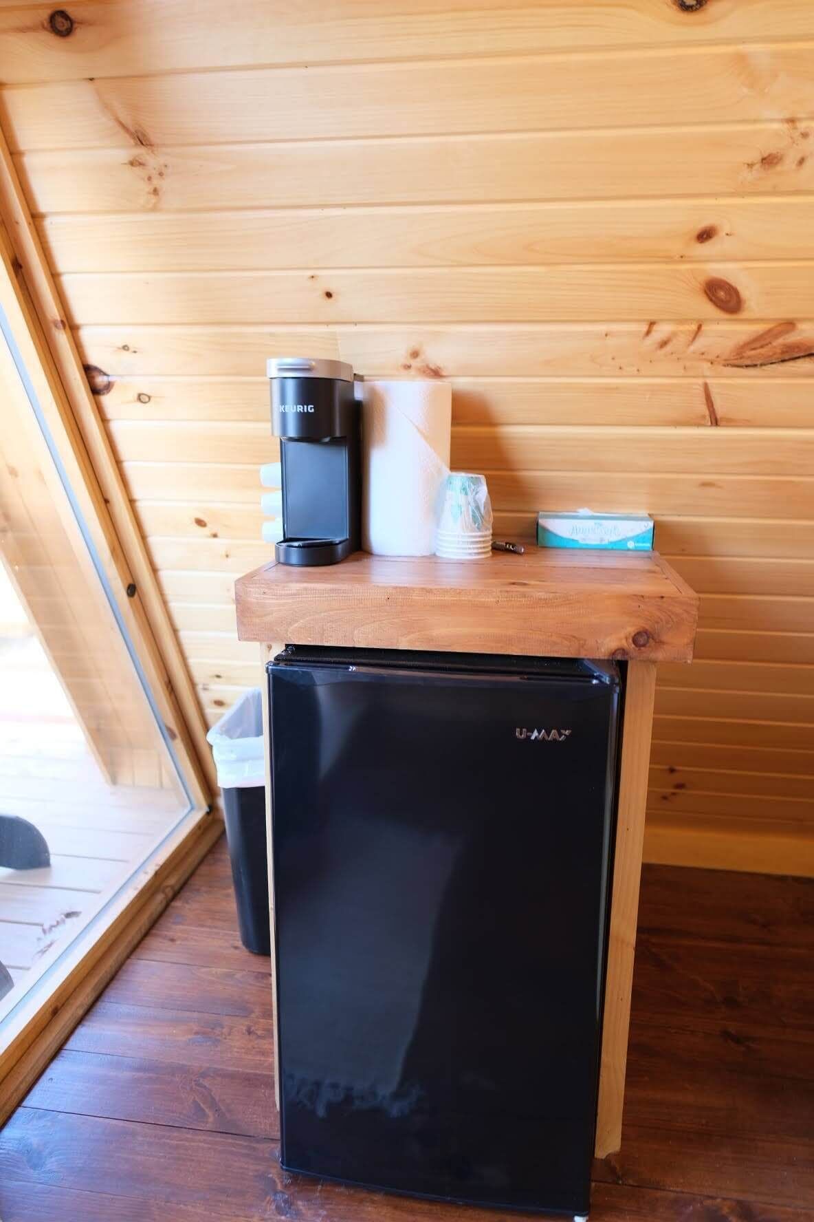 A compact kitchenette with a black U-Matic refrigerator, a Keurig coffee maker, and a stack of paper towels on a wooden countertop.