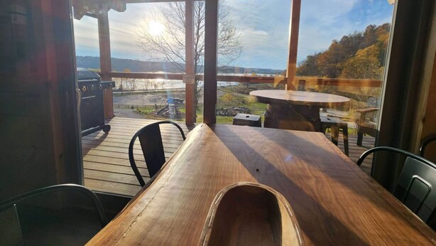 Una terraza de madera con una mesa y sillas, con vistas a un paisaje pintoresco que incluye una masa de agua y colinas.
