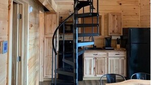 Interior de una cabaña de madera con una escalera en espiral, armarios de madera, una mesa de comedor y un refrigerador negro.