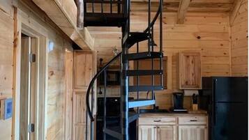 A wooden cabin interior with a spiral staircase, wooden cabinets, a dining table, and a black refrigerator.