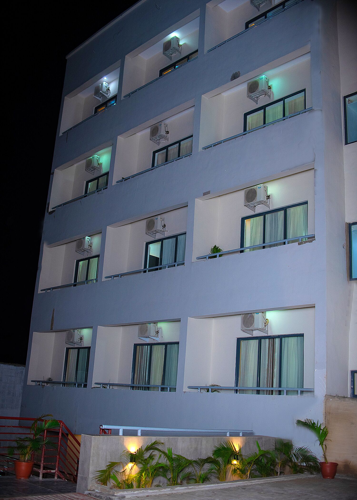 A multi-story hotel with air conditioning units on each window, a red railing, and potted plants at the entrance.