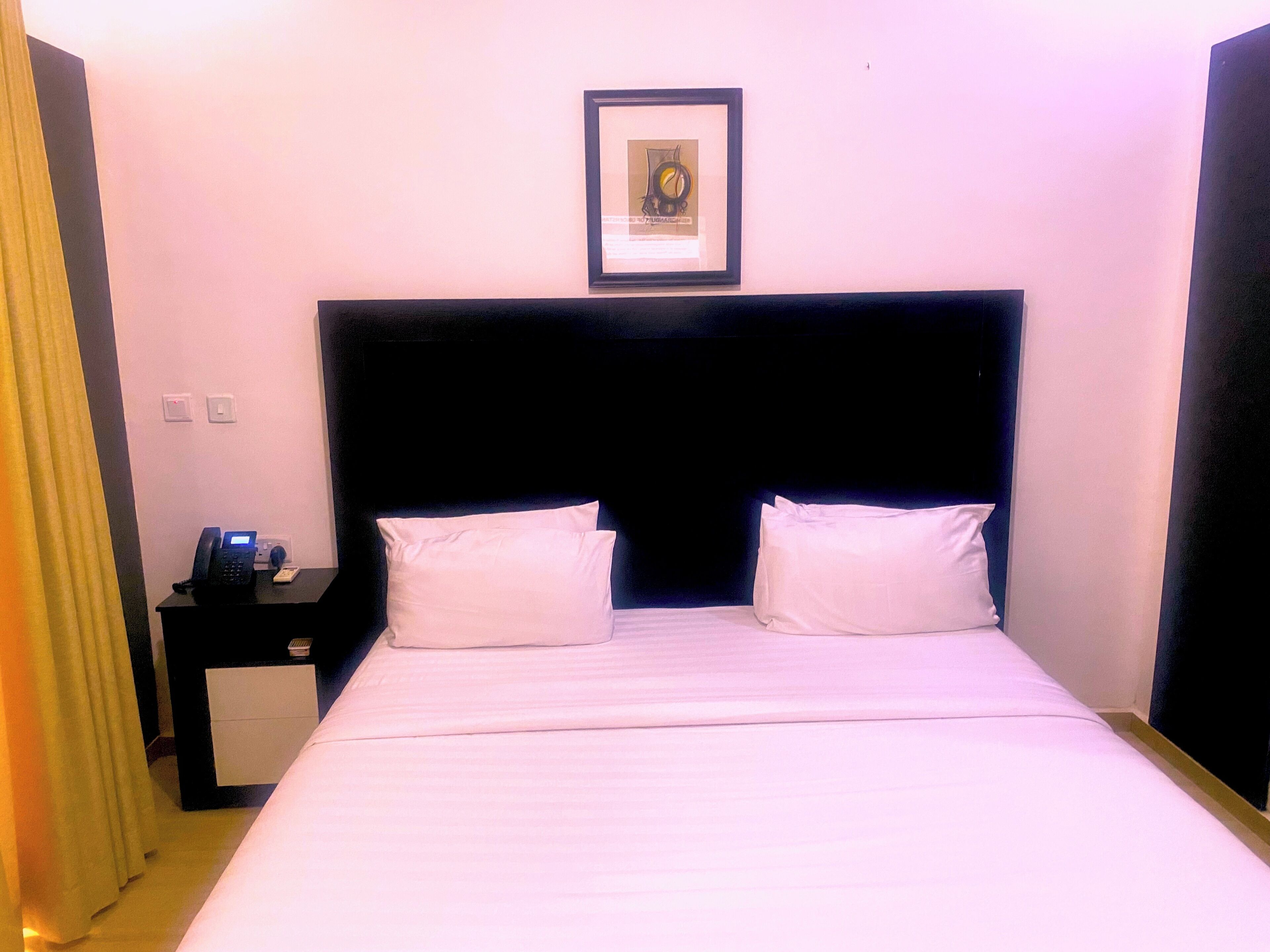 Habitación de hotel con una cama, dos almohadas, una mesita de noche con un teléfono y un cuadro en la pared.
