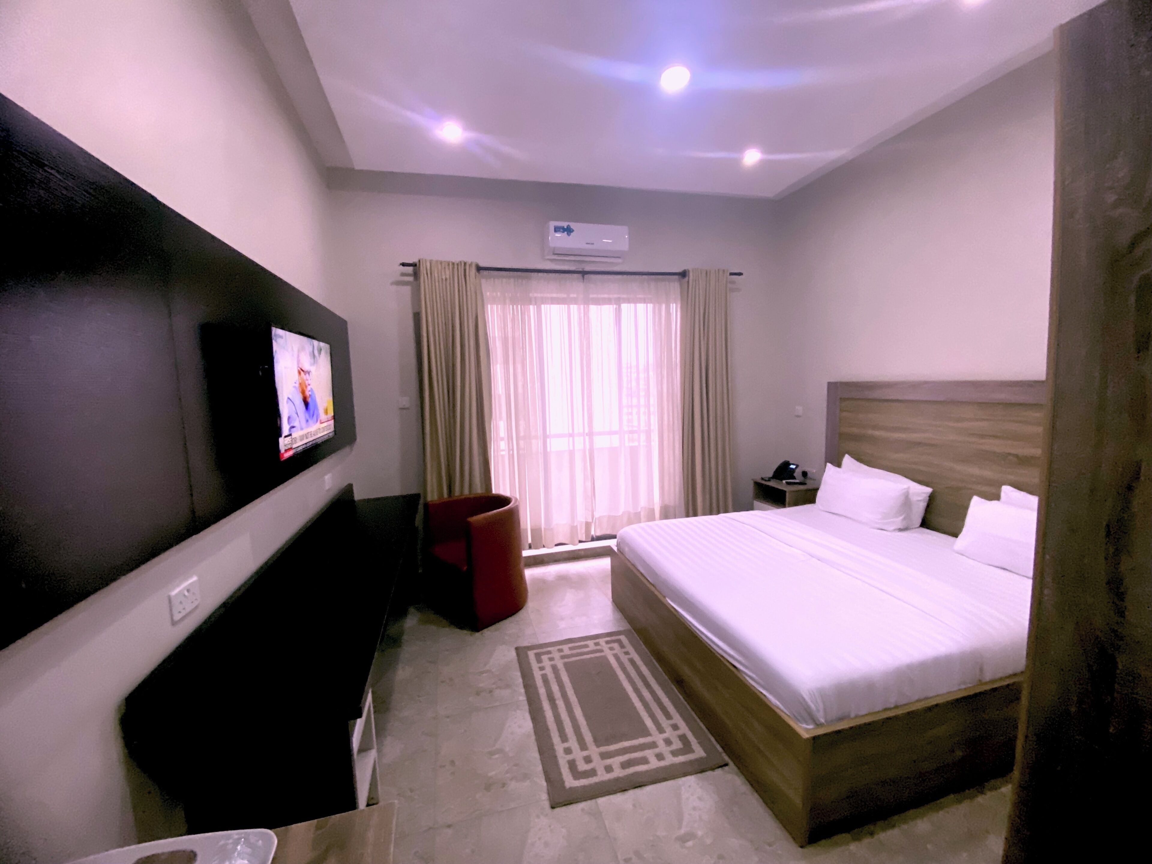 A hotel room with a bed, a TV mounted on the wall, a bedside table with a phone, and a window with curtains.