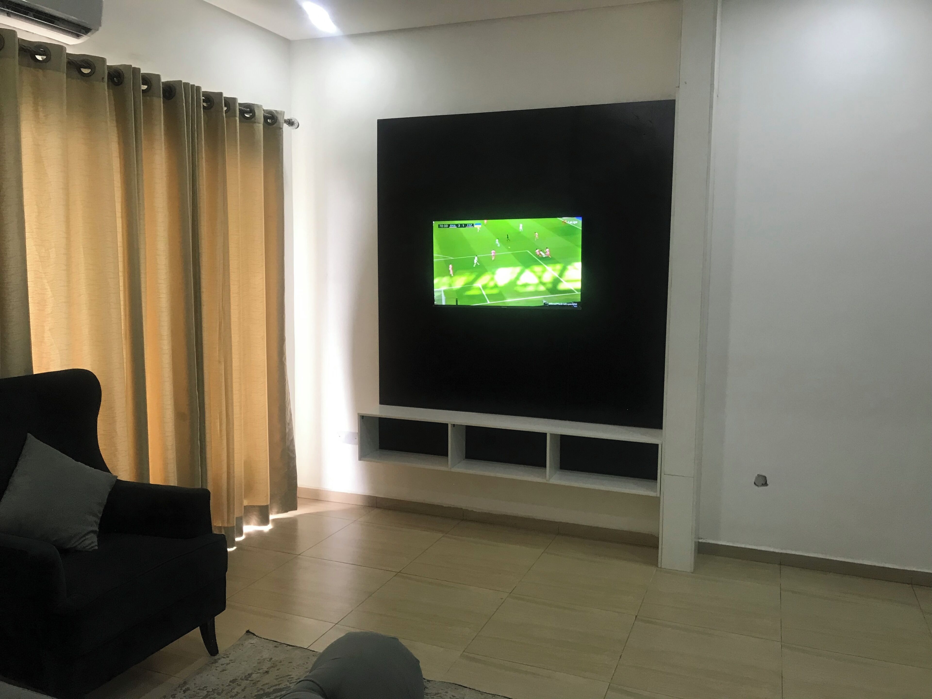 A living room with a flat-screen TV mounted on the wall, a black sofa, and a curtain.
