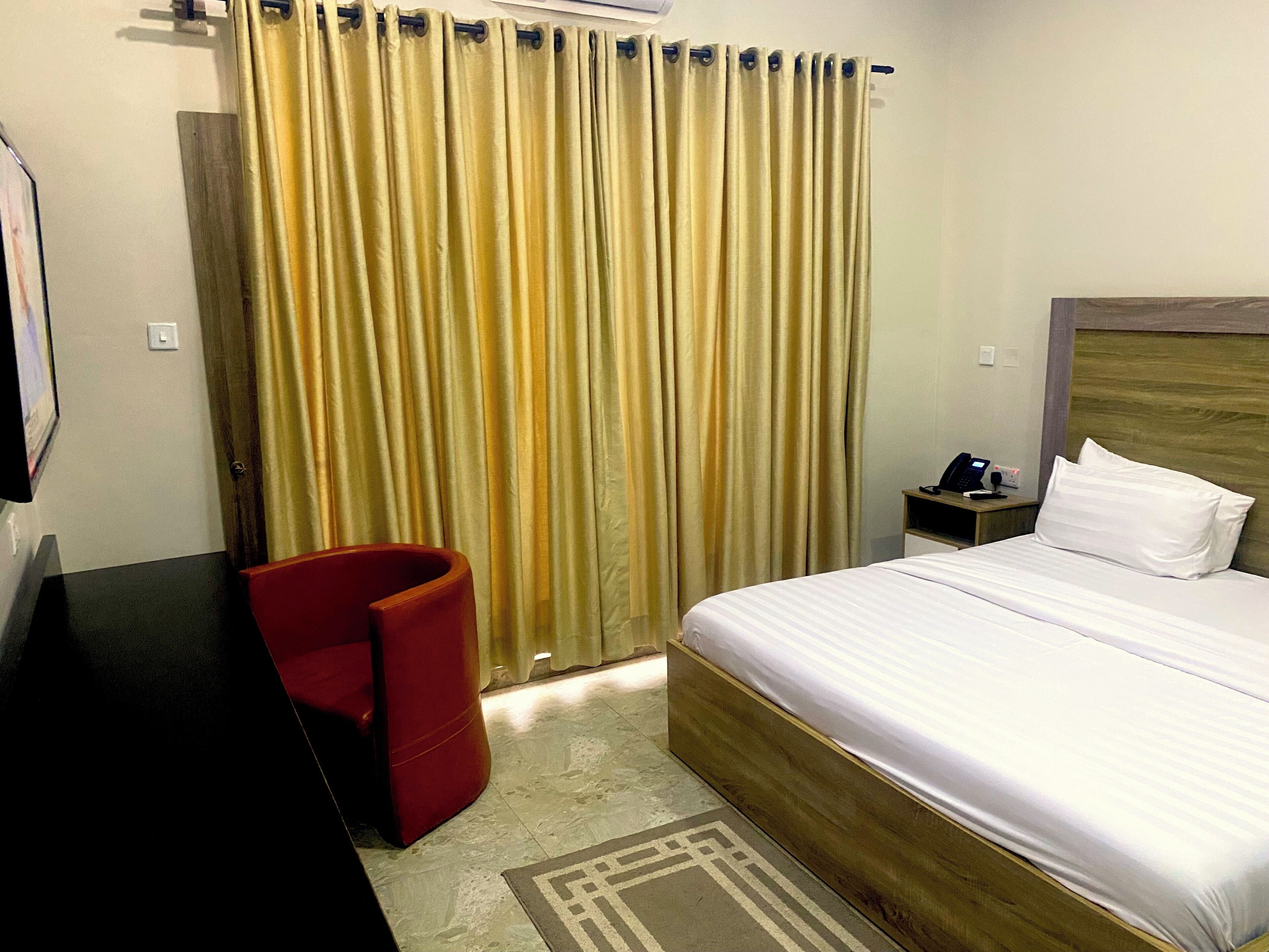 A hotel room with a bed, a desk, a chair, a nightstand, and a curtain.