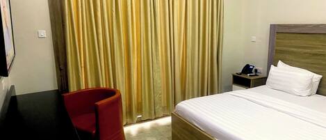 A hotel room with a bed, a desk, a chair, a nightstand, and a curtain.