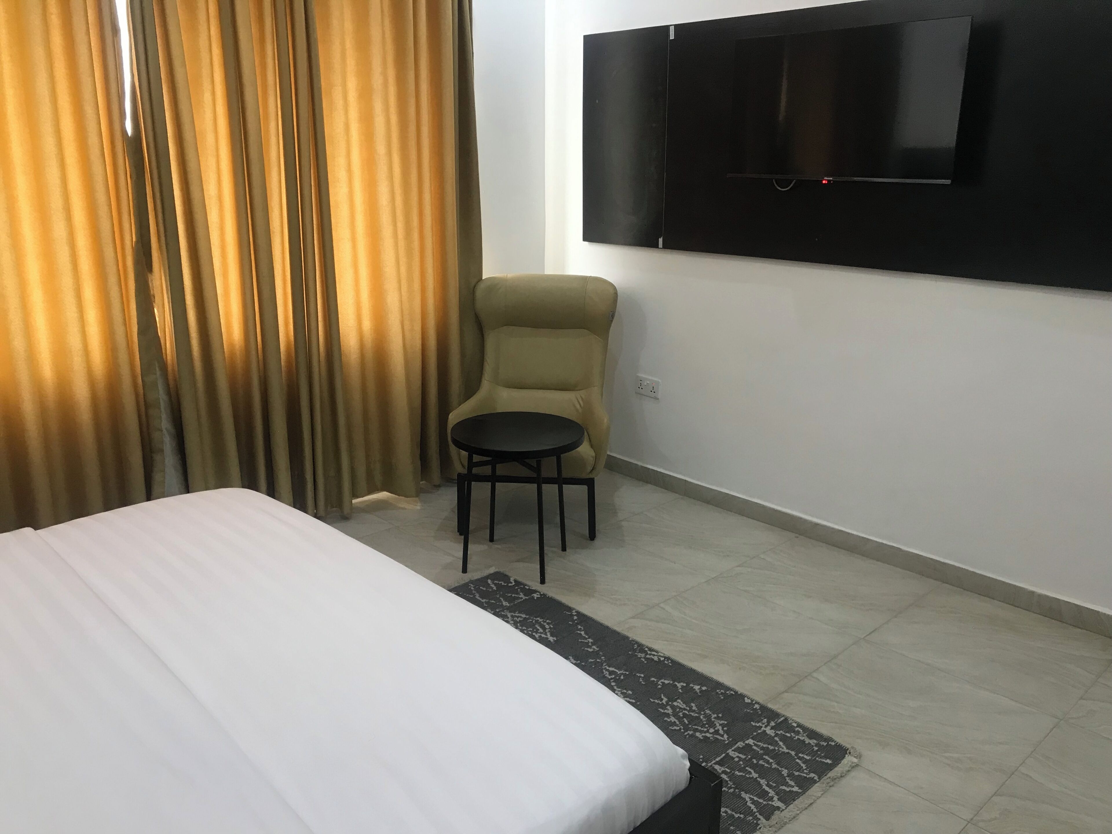 A hotel room with a bed, a small table, a chair, a TV mounted on the wall, and a window with curtains.