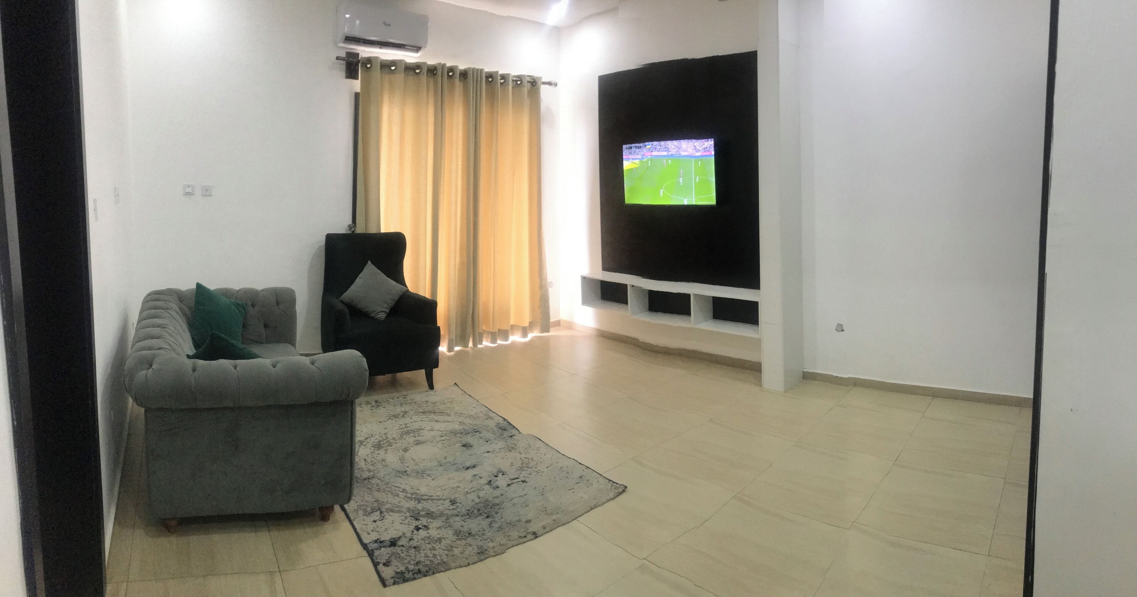 Un salon avec un canapé gris, un fauteuil noir, un grand téléviseur fixé au mur et un rideau beige.