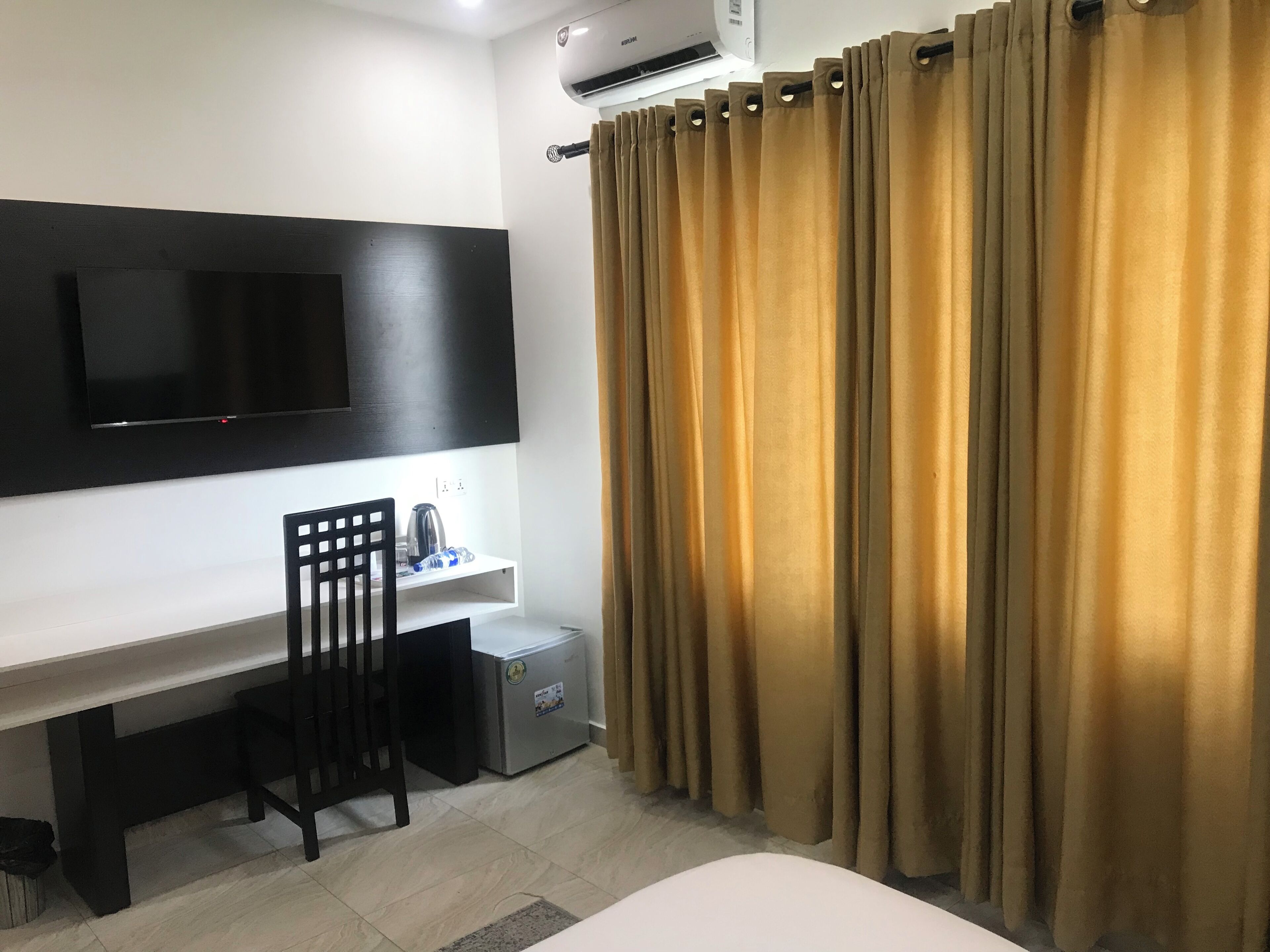 A hotel room with a flat-screen TV mounted on the wall, a desk with a chair, a small refrigerator, and a bed.