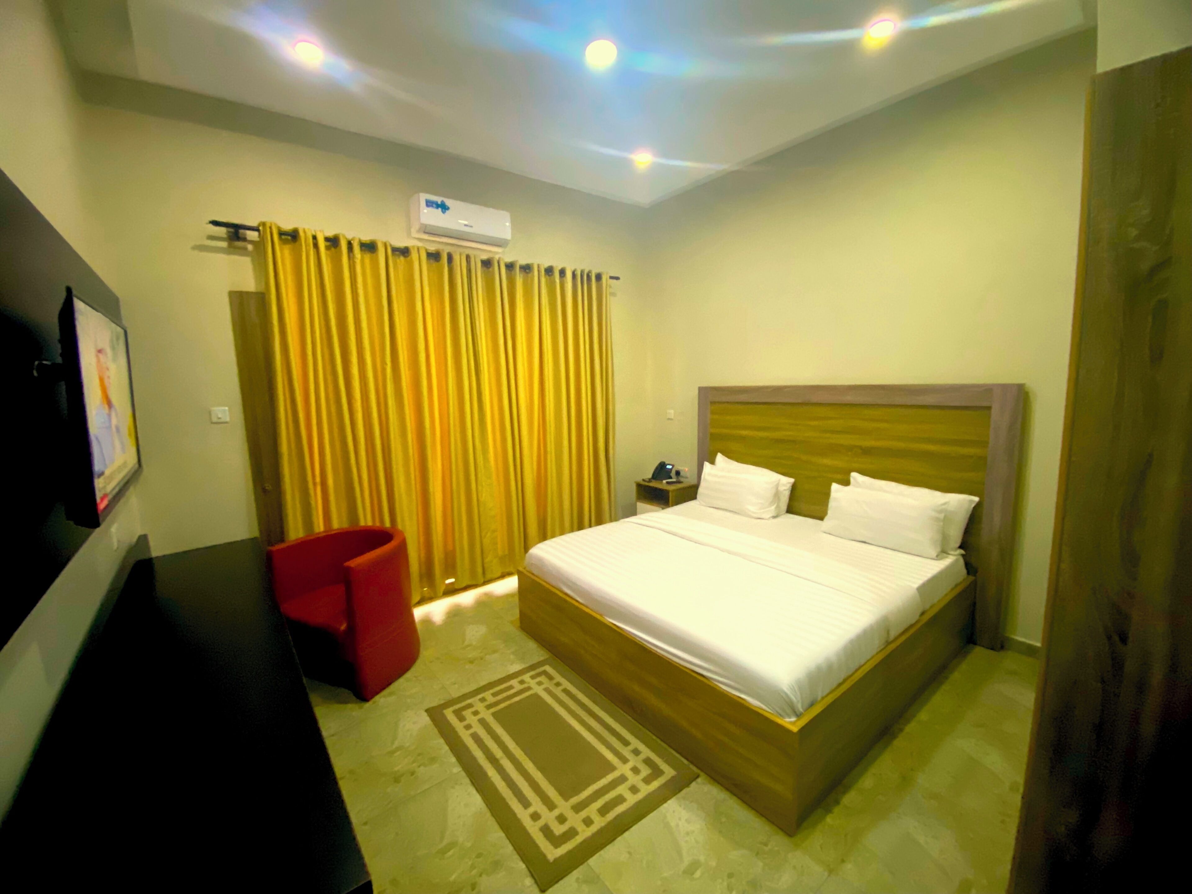 A hotel room with a bed, a TV, a red chair, a bedside table, and a curtain.