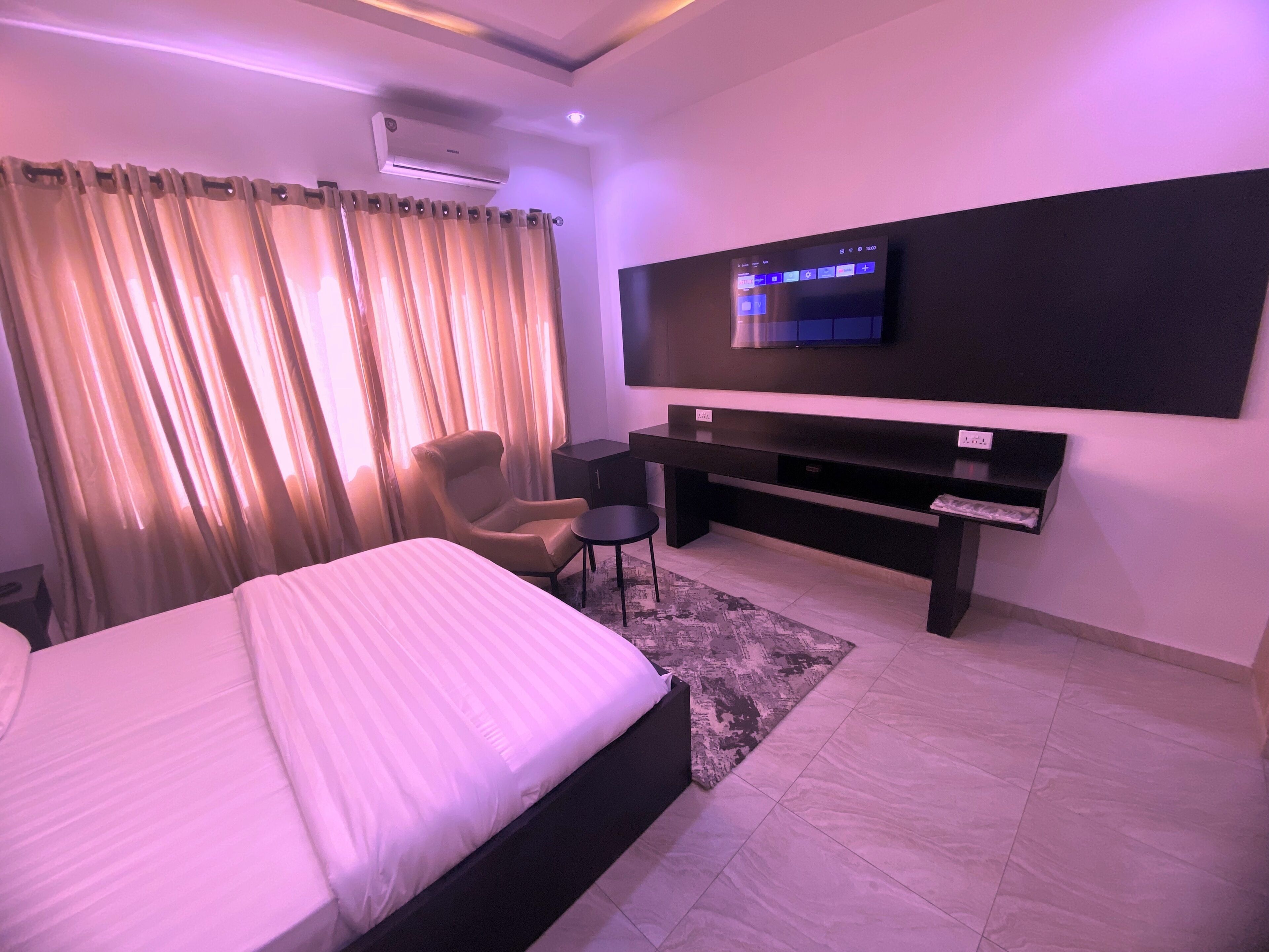 A hotel room with a bed, a desk, a chair, a TV, and an air conditioning unit.
