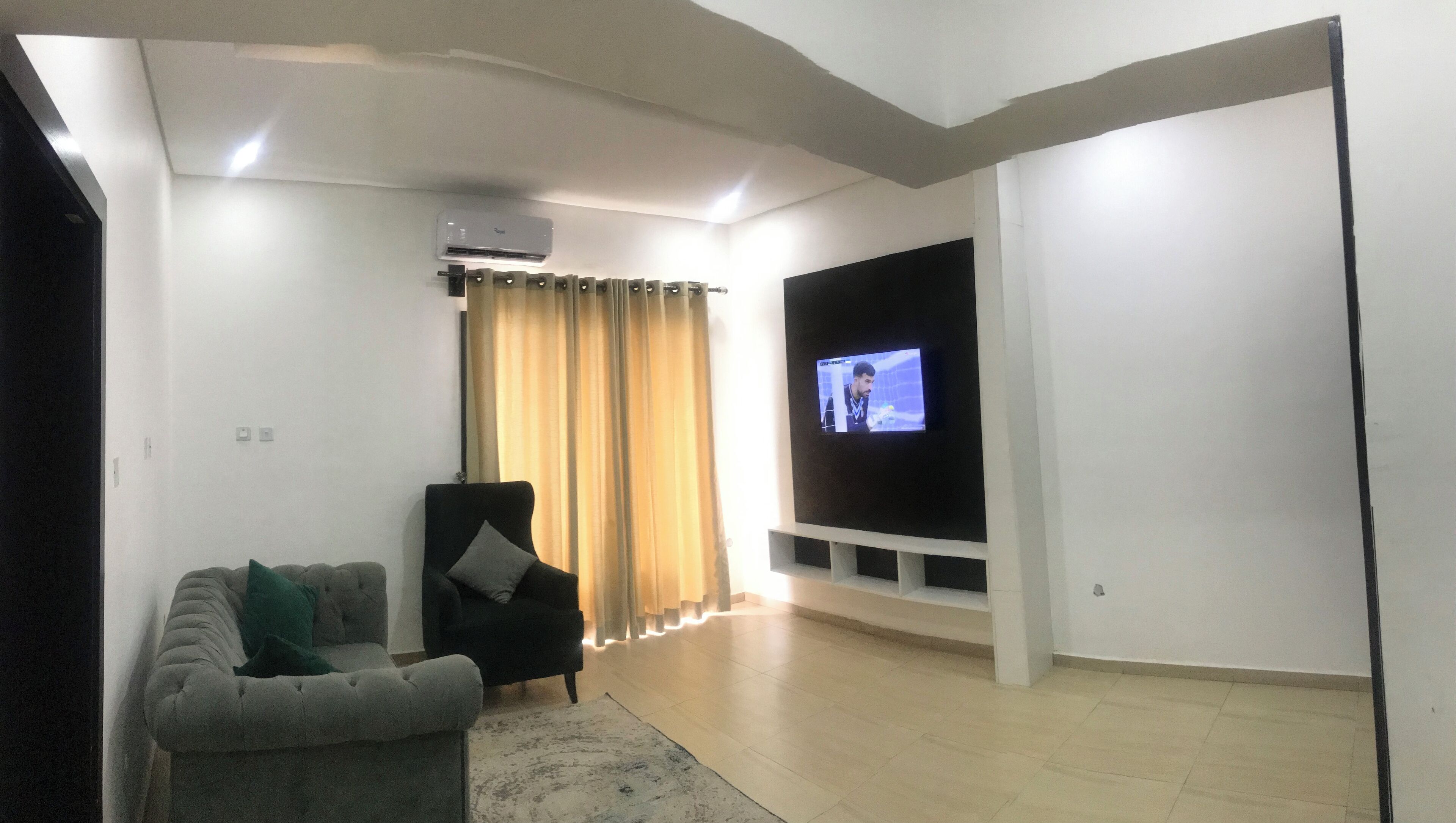 A living room with a grey sofa, a black armchair, a flat-screen TV mounted on a white wall, and an air conditioning unit on the ceiling.