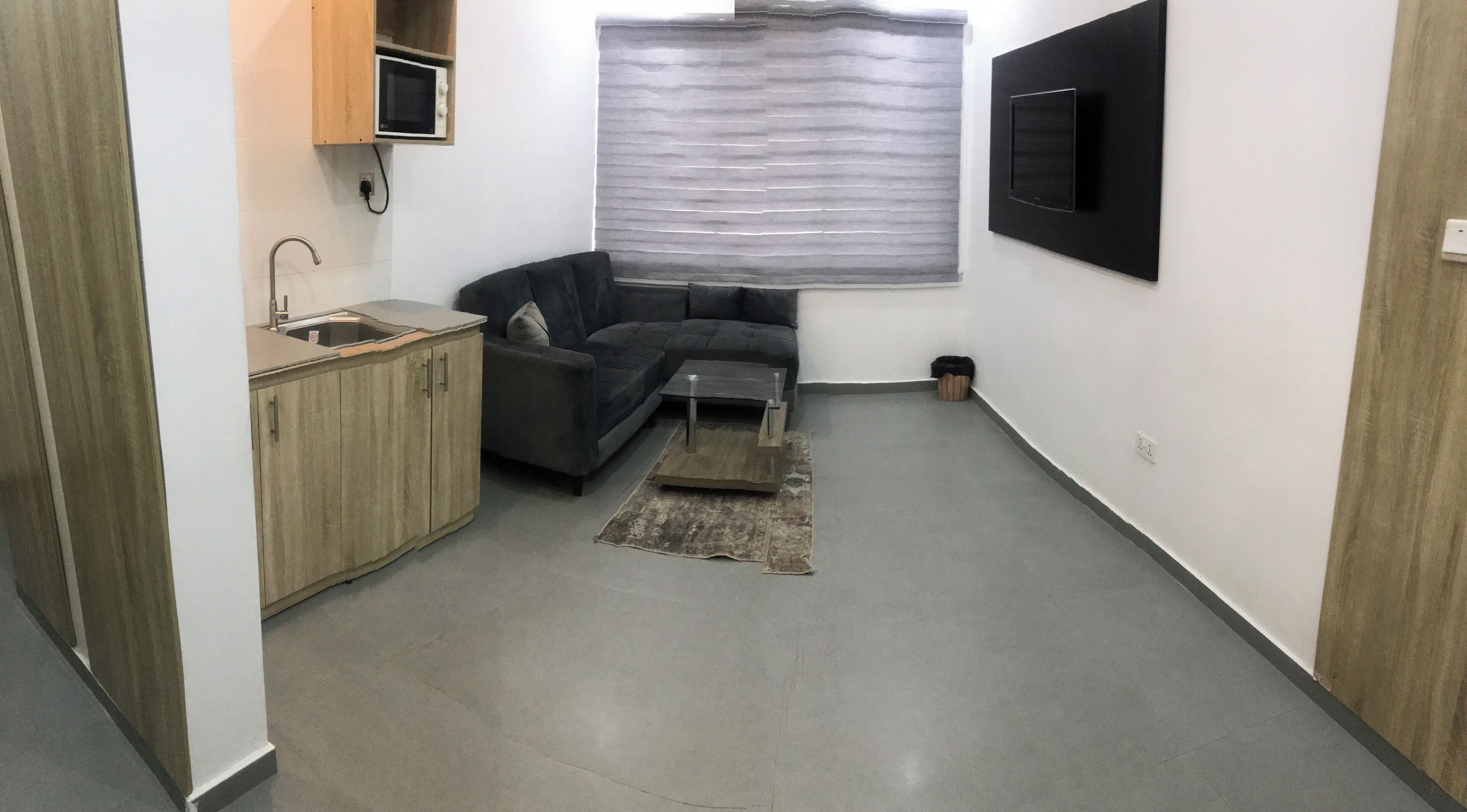 Een ruimtelijk ingerichte leefruimte met kitchenette, een bank, een klein bijzettafeltje en een aan de muur bevestigde televisie.
