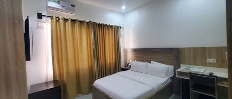 A hotel room with a bed, a bedside table with a phone, a small desk with a cup and a remote control, and a curtain.