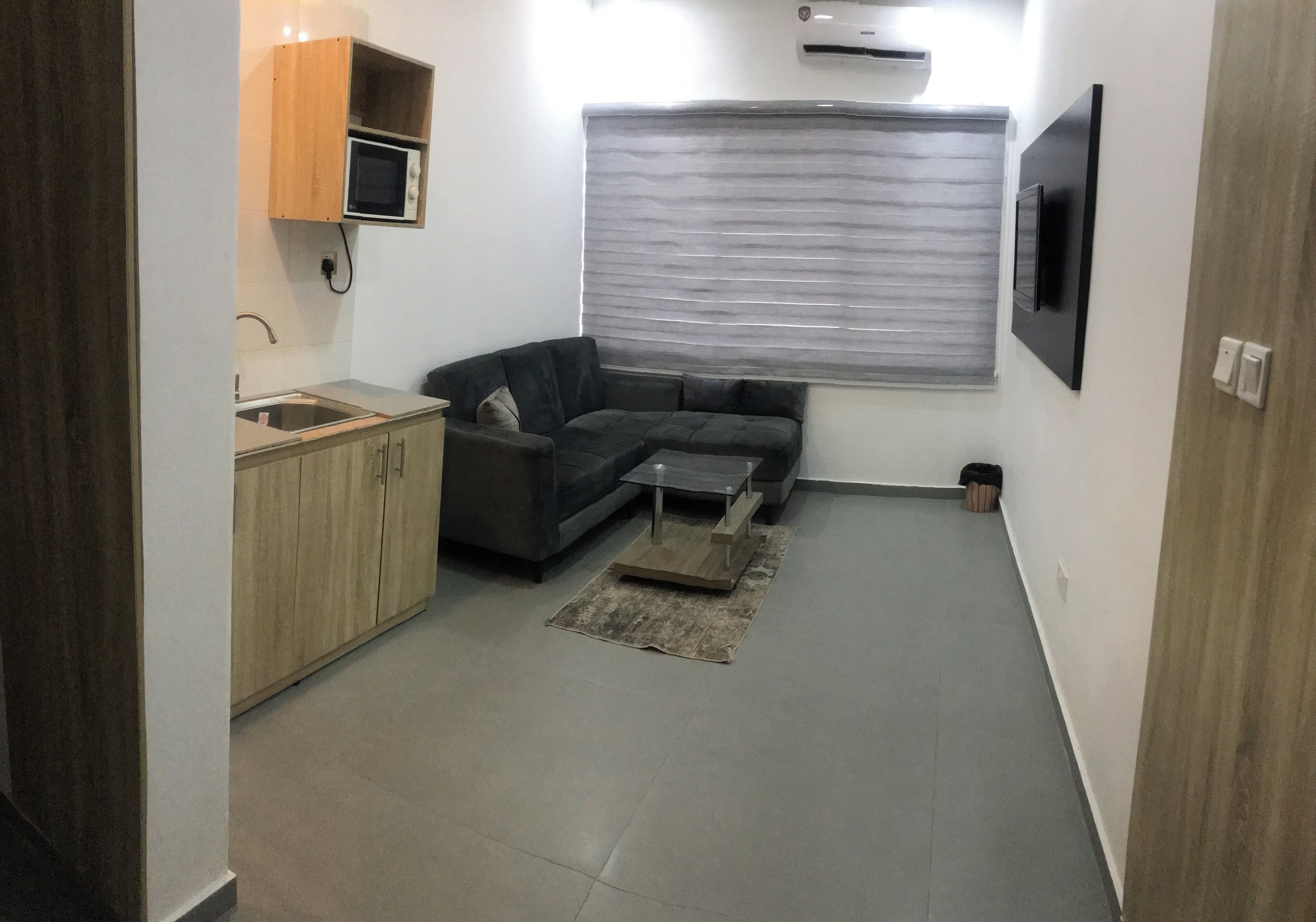 A compact living space with a kitchenette, a gray sofa, a small coffee table, and a window with blinds.