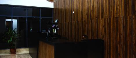 Egy recepciós pult, melynek háttérfalát fából készült panelek borítják, egy virágtartóban lévő növény és egy üvegajtó látható a háttérben.