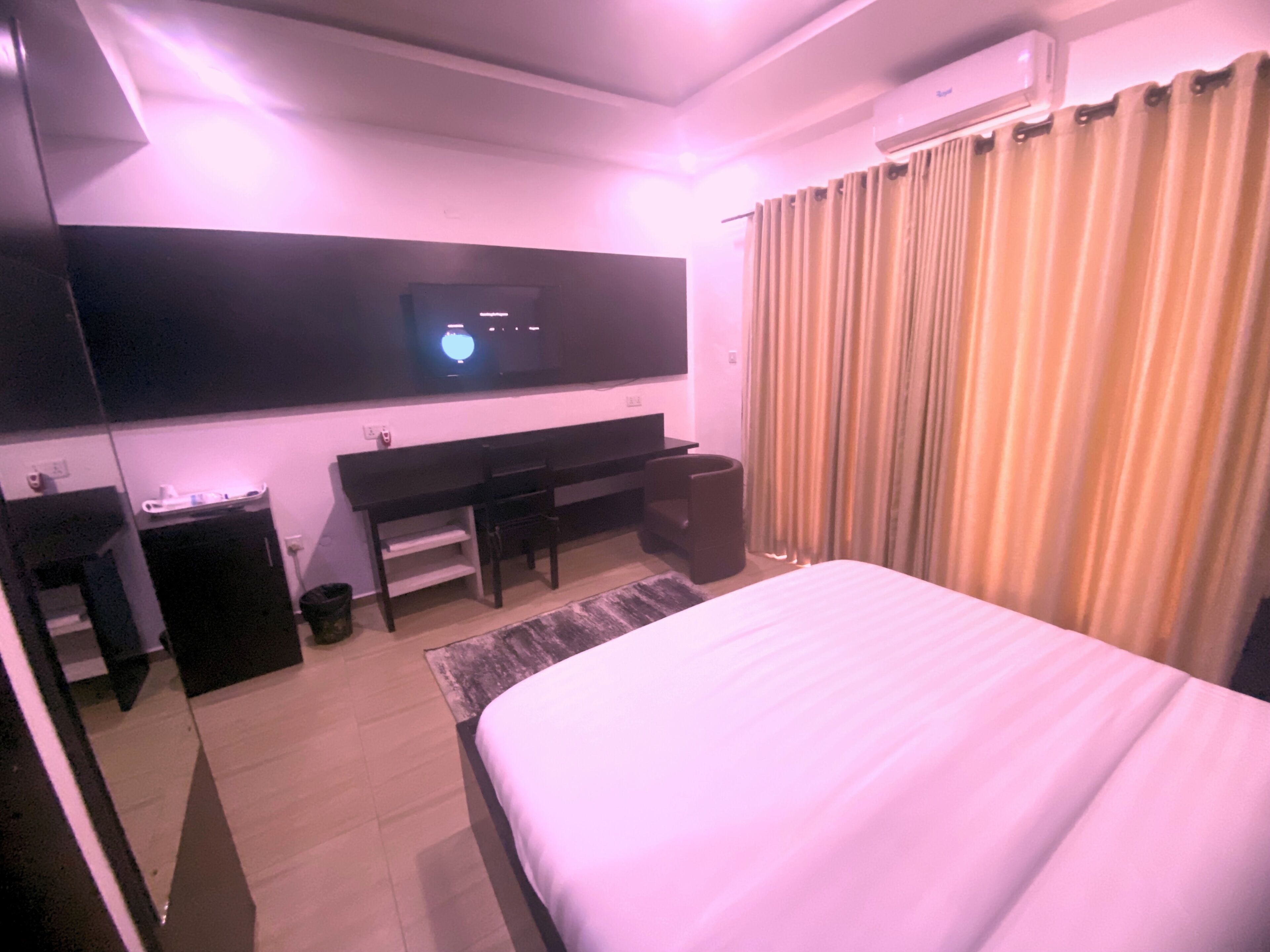 A hotel room with a bed, a desk, a chair, a TV mounted on the wall, and a window with curtains.