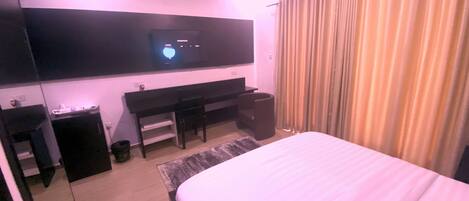 A hotel room with a bed, a desk, a chair, a TV mounted on the wall, and a window with curtains.