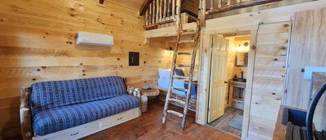 Ein gemütliches Blockhaus-Interieur mit Holzboden, einer Couch, einer Leiter und einem Schlafzimmer auf der Empore mit Holzwand.