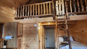 Interior rústico de una cabaña con un dormitorio en entrepiso, paredes de madera, un ventilador de techo y una zona de cocina.
