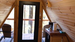 Quarto sótão aconchegante com teto inclinado de madeira, uma cama, uma mesinha com abajur e janela com vista para um deck.