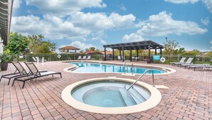 Piscina con sillas de descanso y jacuzzi, rodeada de un patio de ladrillo y una zona de estar cubierta.