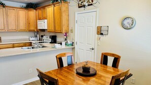A kitchen with wooden cabinets, a white door, a dining table with chairs, a chandelier, and a wall clock.