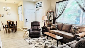 A living room with a brown leather sofa, a matching armchair, a wooden coffee table, and a patterned rug. There is a dining table with chairs, a small side table with a lamp, and a cabinet with glass doors.