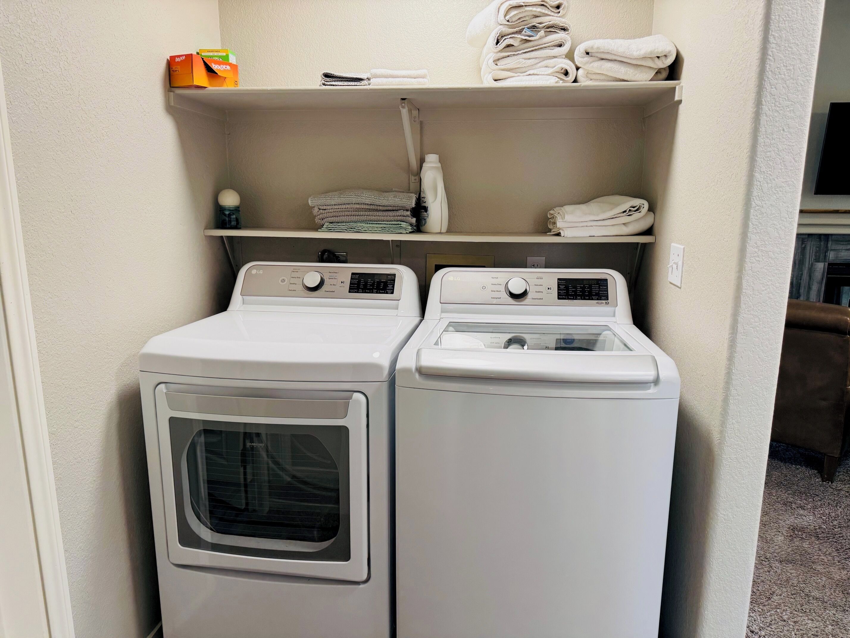 A laundry area with two white front-loading washing machines and a shelf above containing folded towels and a box of laundry detergent.