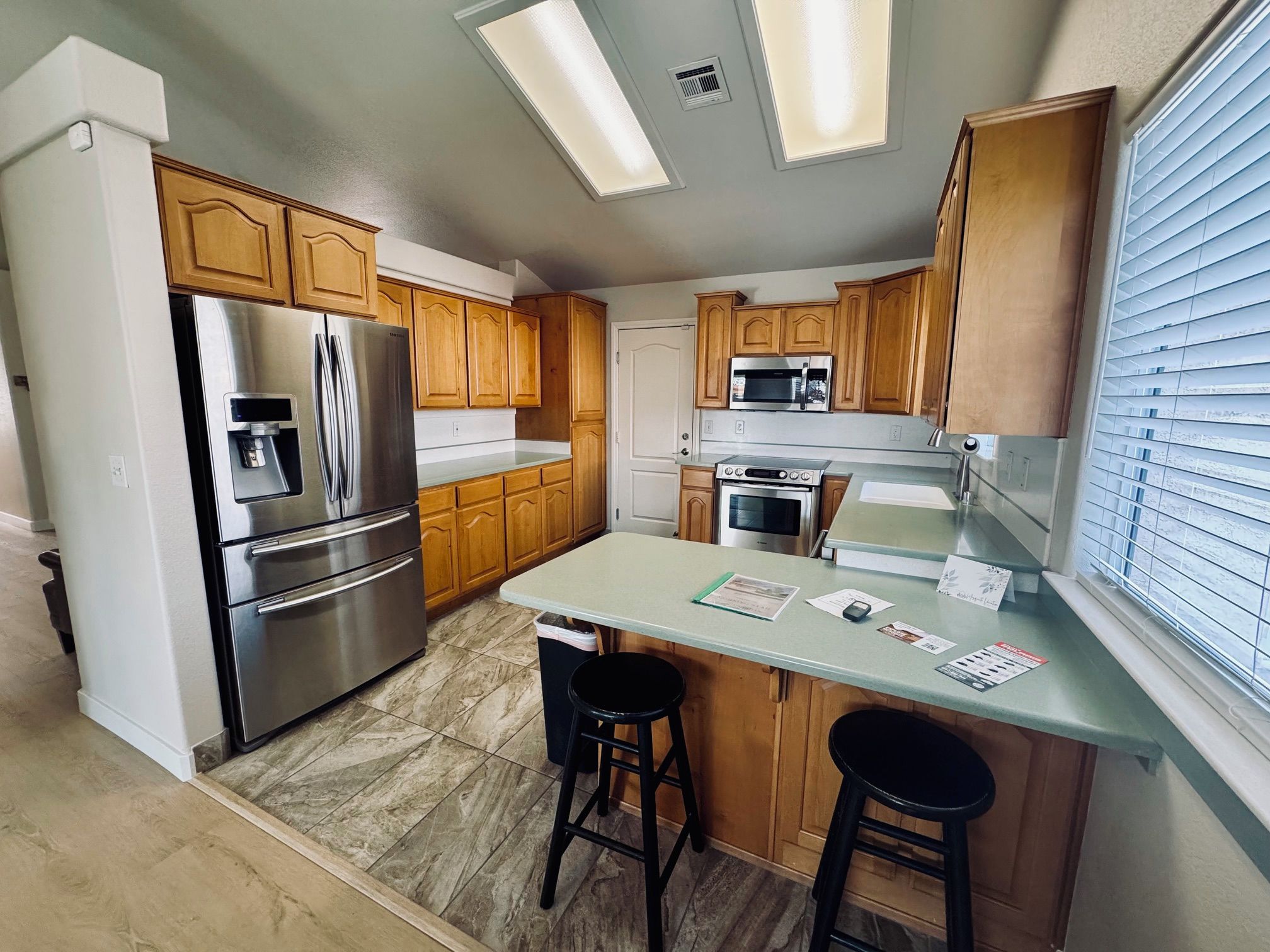 A modern kitchen with stainless steel appliances, wooden cabinets, a central island with stools, and a window with blinds.
