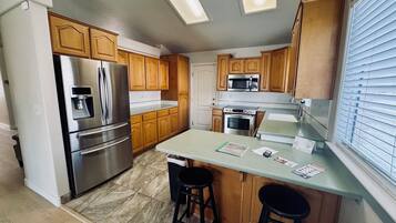 A modern kitchen with stainless steel appliances, wooden cabinets, a central island with stools, and a window with blinds.