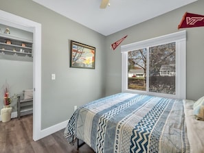Quarto com uma janela grande, uma cama com manta estampada, um quadro na parede e uma estante com objetos decorativos.
