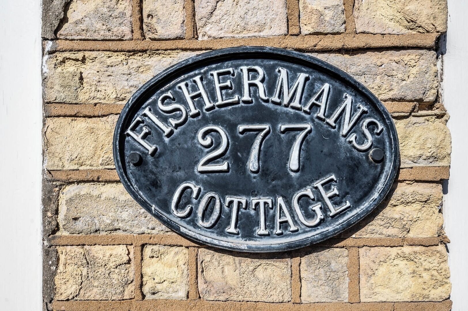 A metal plaque with 'FISHERMANS 277 COTTAGE' engraved on it, mounted on a brick wall.