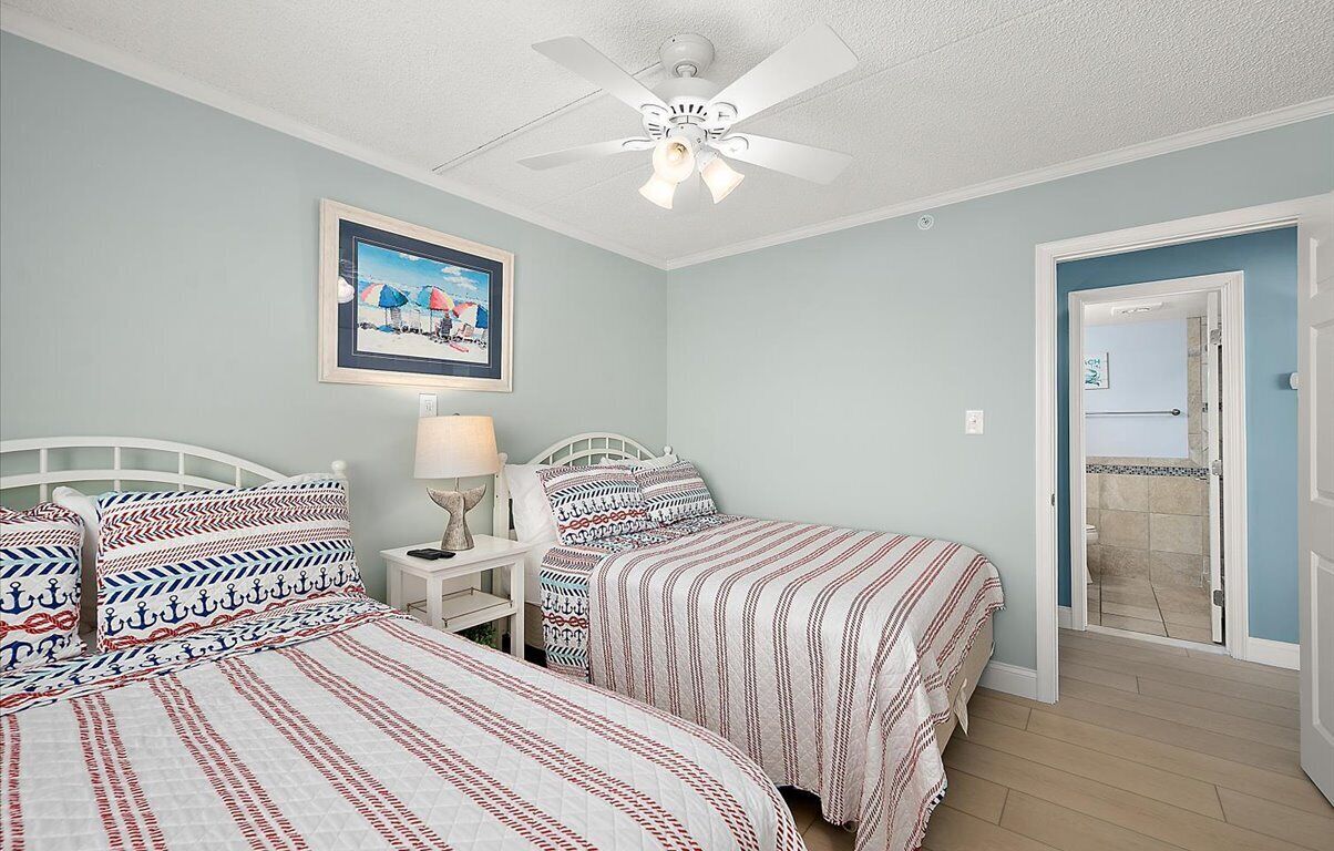 A bedroom with two beds, a ceiling fan, a nightstand with a lamp, a framed picture on the wall, and a bathroom door in the background.