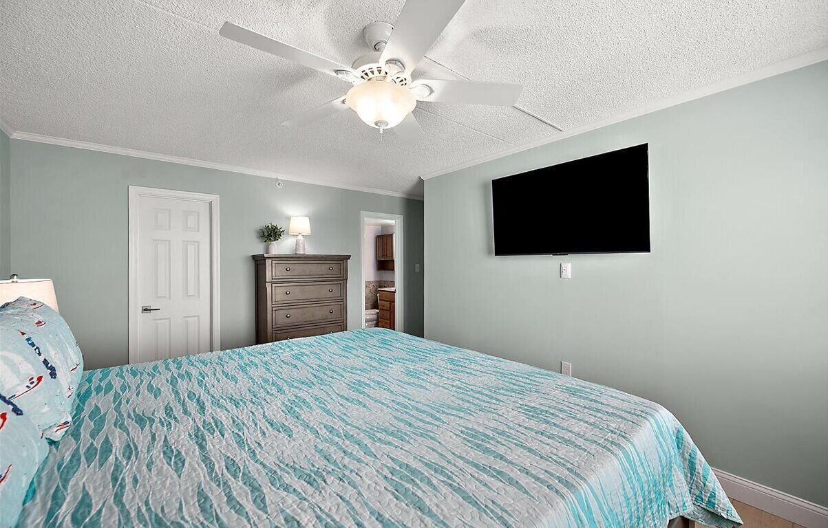 A bedroom with a bed, a dresser, a ceiling fan, and a TV mounted on the wall.