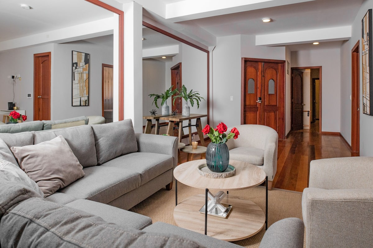 A spacious living area with a grey sofa, a round coffee table, and a vase with red flowers. There is a wooden dining table with chairs and a small table with decorative items.