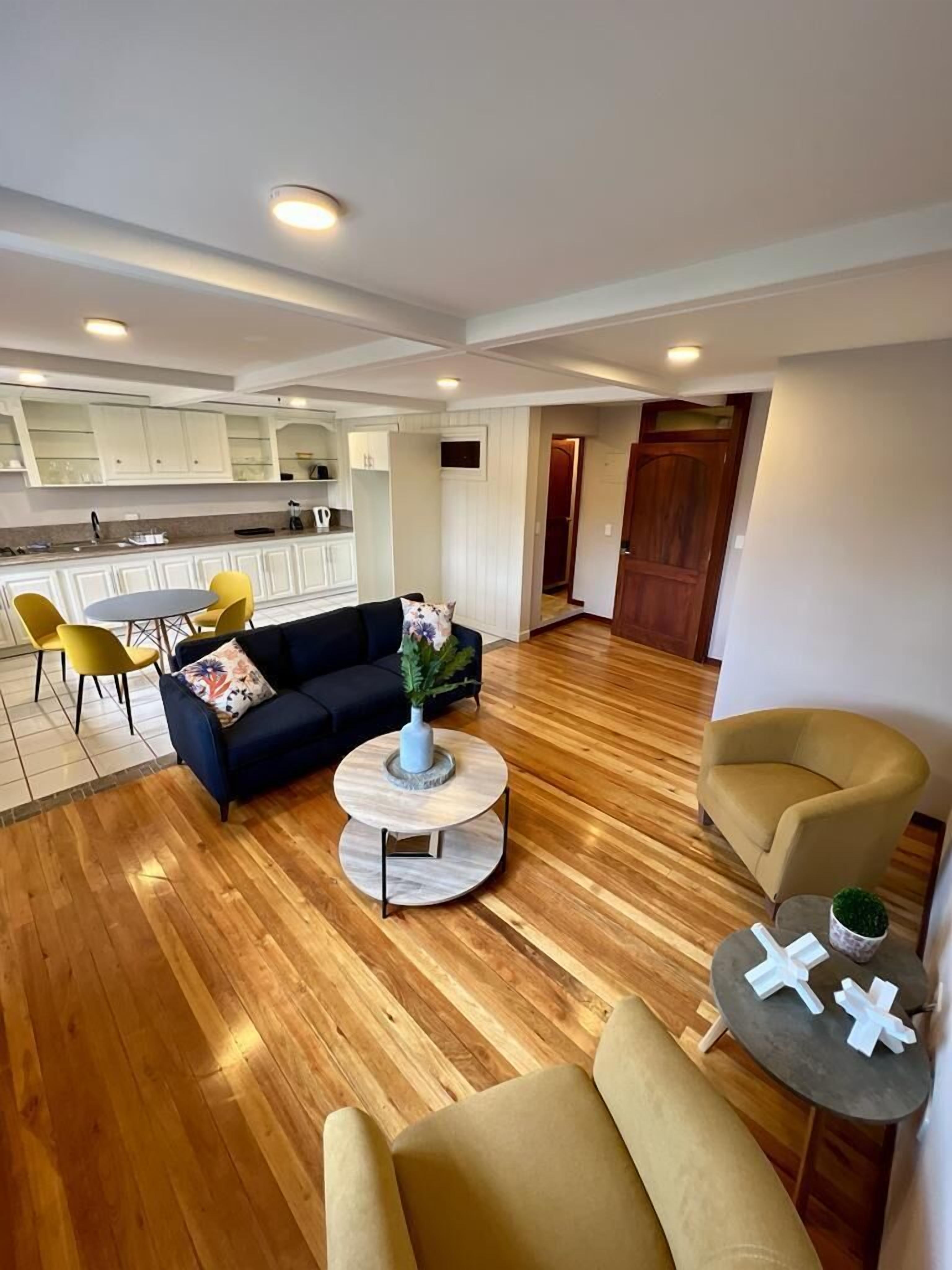 A spacious living area with a sofa, two armchairs, a coffee table, and a dining table with yellow chairs. A kitchen with white cabinets and a wooden floor is visible in the background.
