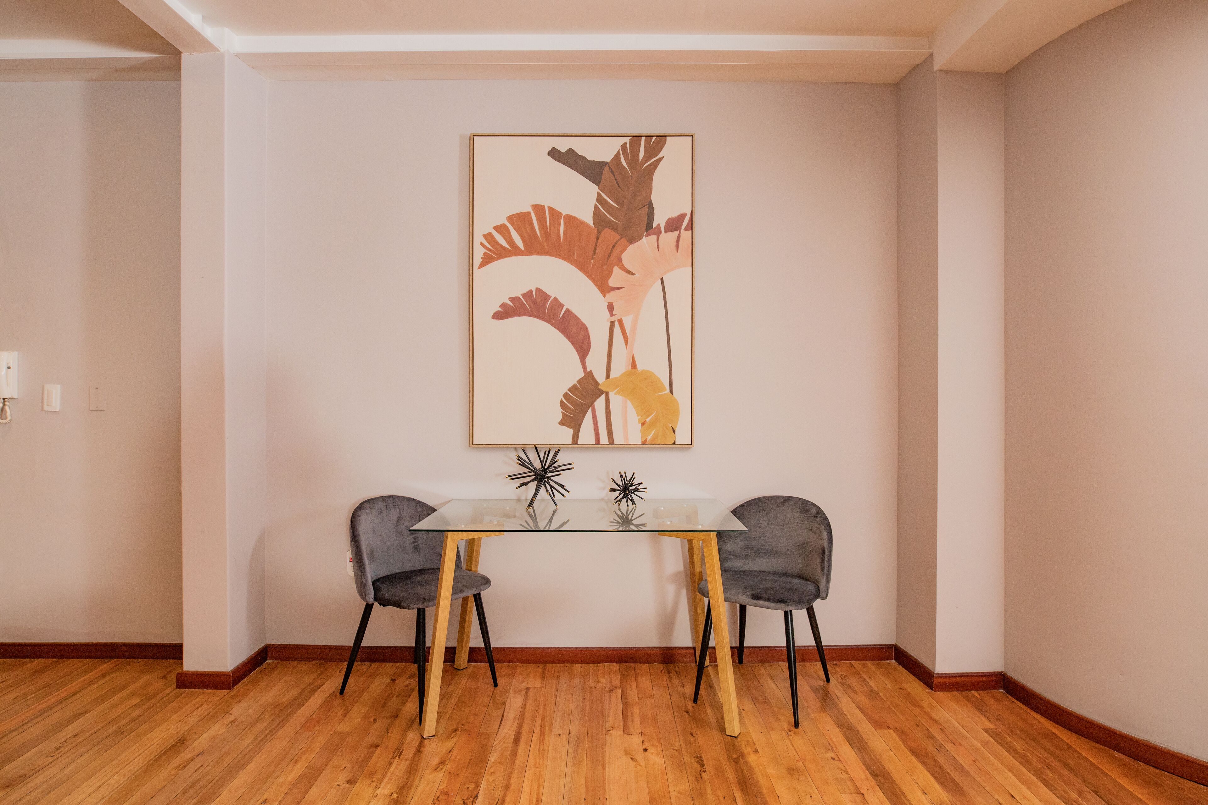 A dining area with a glass table and two chairs, a framed artwork on the wall, and wooden flooring.