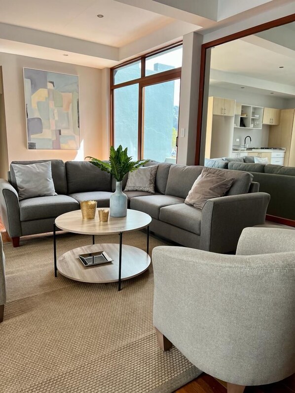 Un salón moderno con un sofá gris, una mesa de centro redonda y un ventanal con vistas a la cocina.