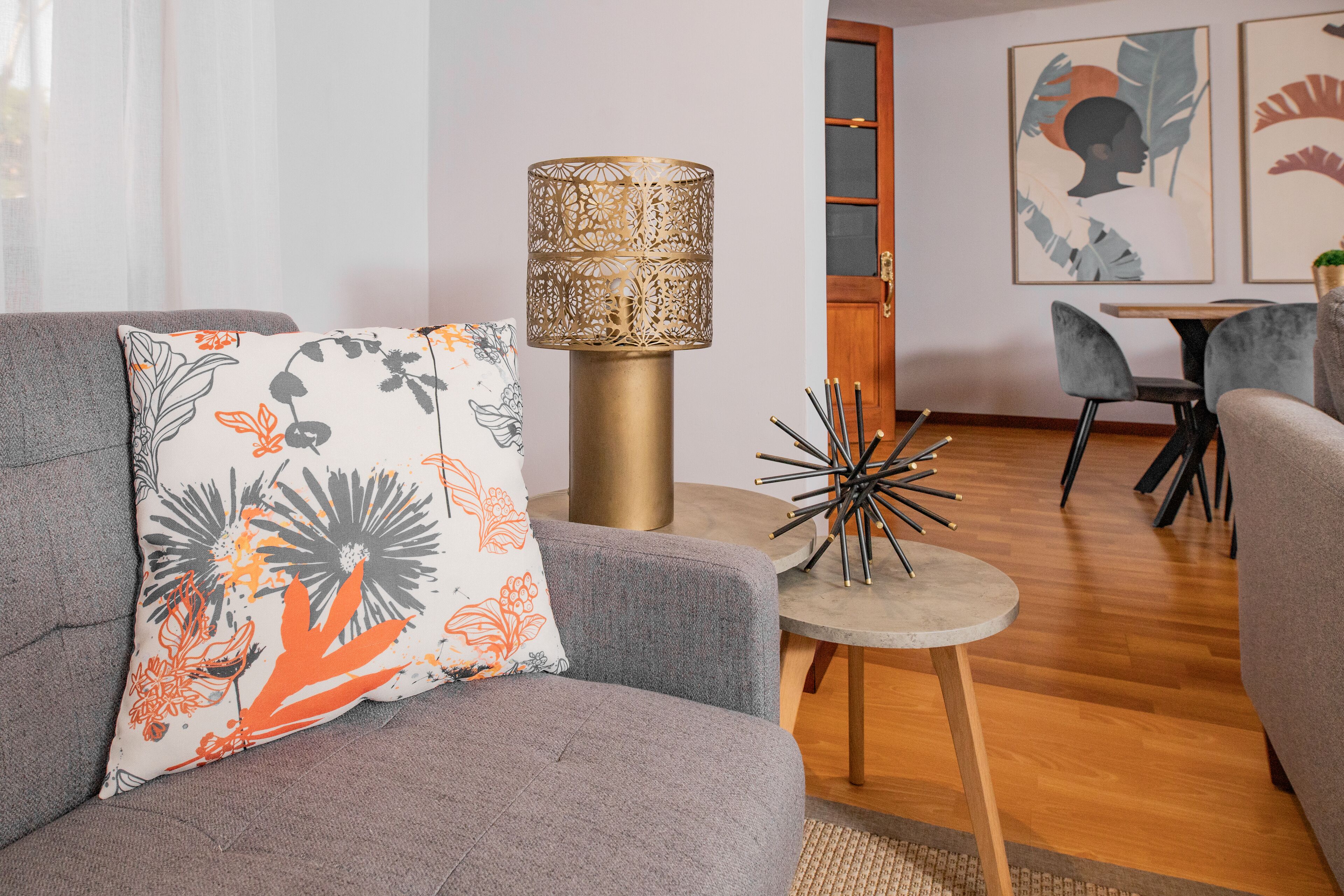 Ein modernes Wohnzimmer mit grauem Sofa, einem gemusterten Zierkissen, einer dekorativen Lampe und einem Essbereich mit Tisch und Stühlen.