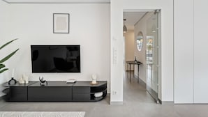A modern living room with a flat-screen TV mounted on the wall, a black entertainment unit, and a glass door leading to another room.