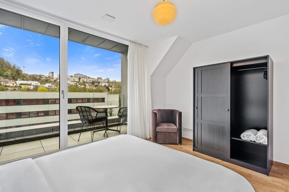 Et soveværelse med en stor seng, et skab, en stol og en balkon med udsigt over byen.