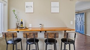 A kitchen with a wooden island, four black bar stools, two framed signs reading "BOX" and "COFFEE", and a plant on the left.